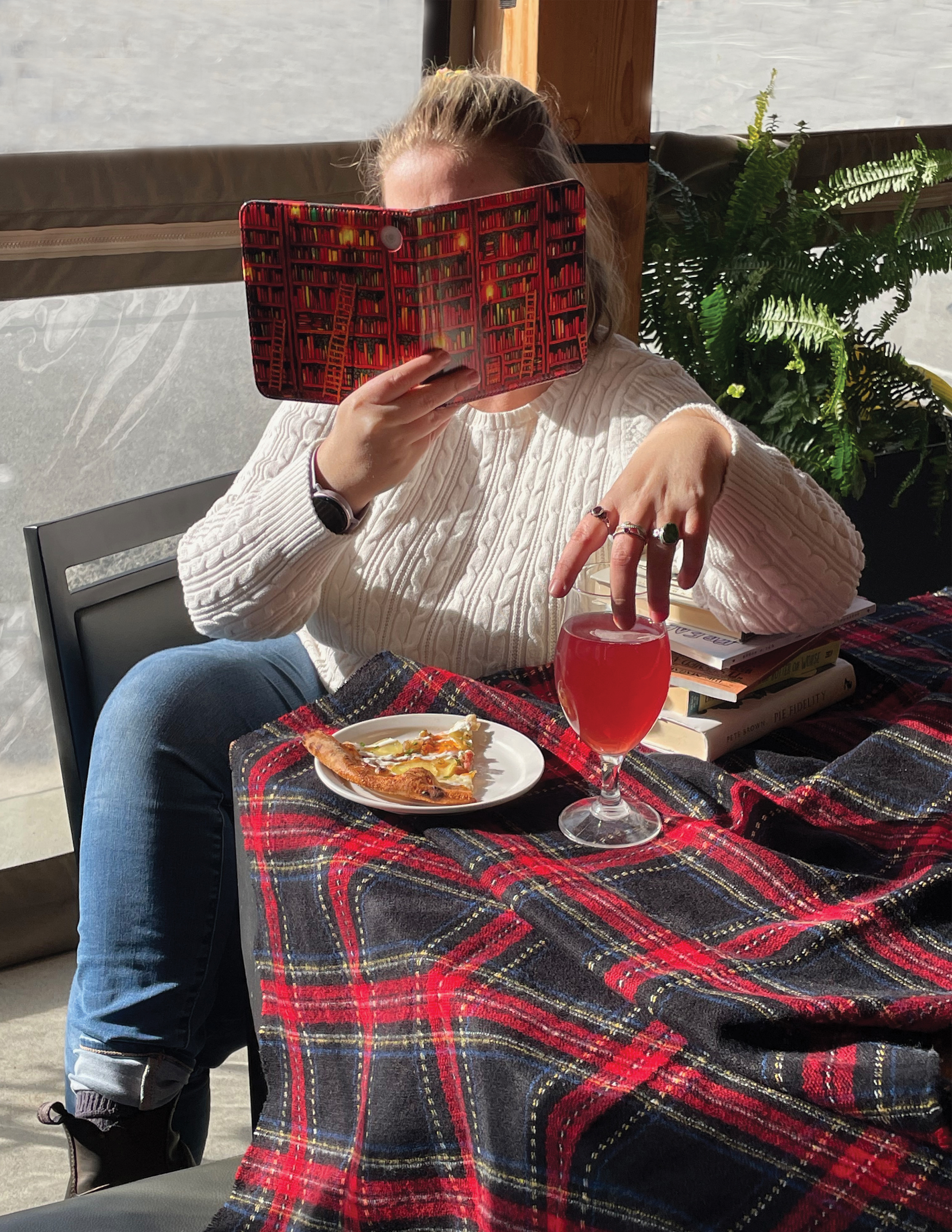 Portrait of Ceilidh, sitting on a restaurant patio reading with a stack of books, food and drink, with their face buried in an e-reader